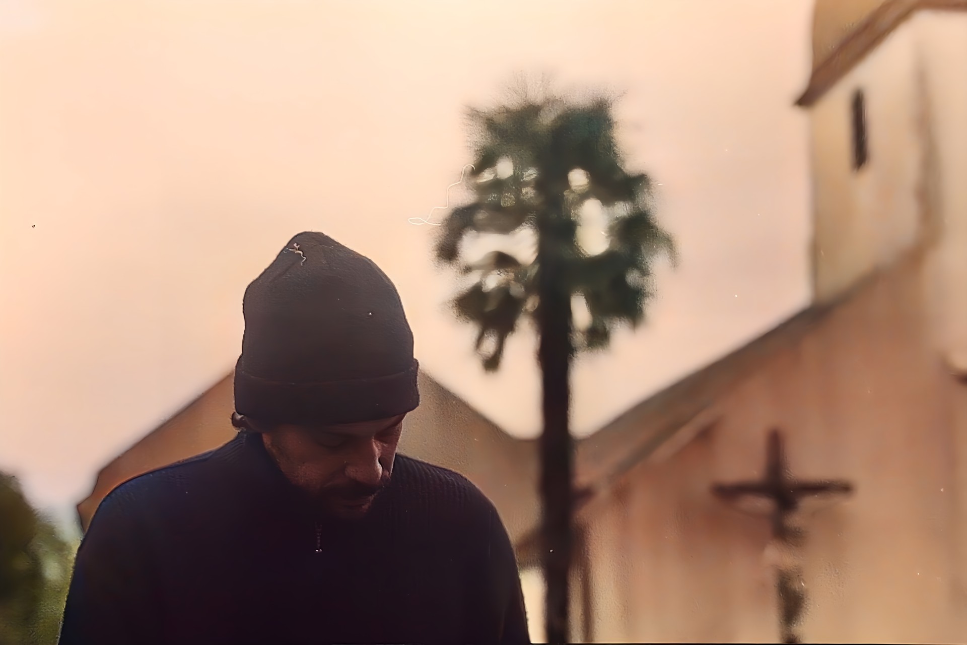 Homme portant un bonnet regardant vers le bas devant une église floue et un palmier, ambiance sépia.